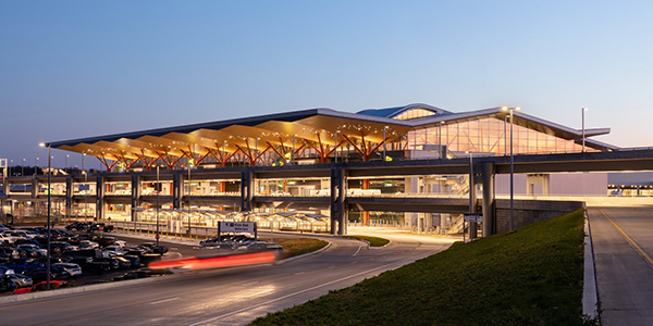 Exterior view of Pittsburgh International Airport at dusk, showing the illuminated terminal with a sweeping roof canopy, glass façade, elevated roadways, parking areas, and vehicle drop-off lanes.