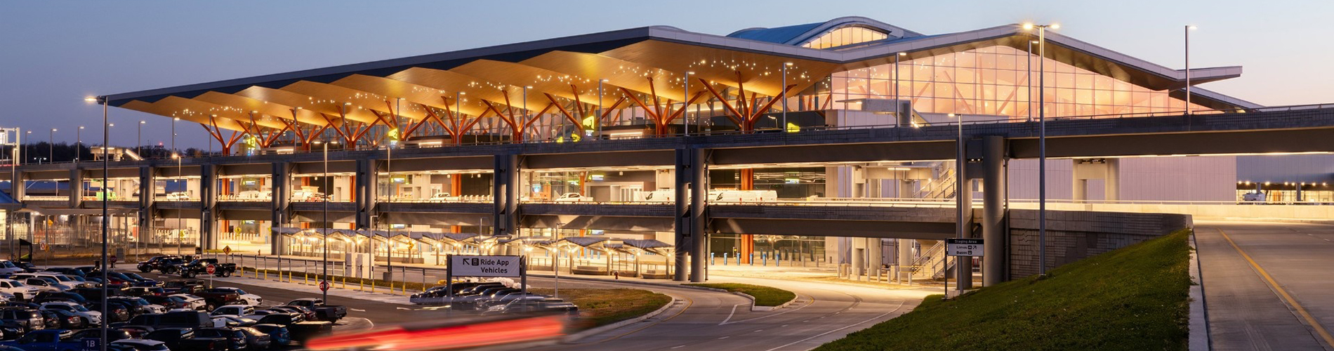 Exterior view of Pittsburgh International Airport at dusk, showing the illuminated terminal with a sweeping roof canopy, glass façade, elevated roadways, parking areas, and vehicle drop-off lanes.