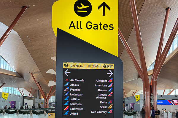 Large yellow “All Gates” airport wayfinding sign with airline listings inside a modern terminal.